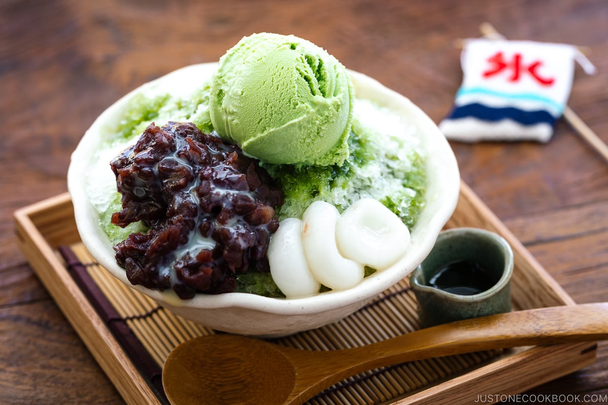 A bowl containing shaved ice, drizzled with matcha syrup and topped with red bean paste, shiratama dango, sweetened condensed milk, and green tea ice cream.