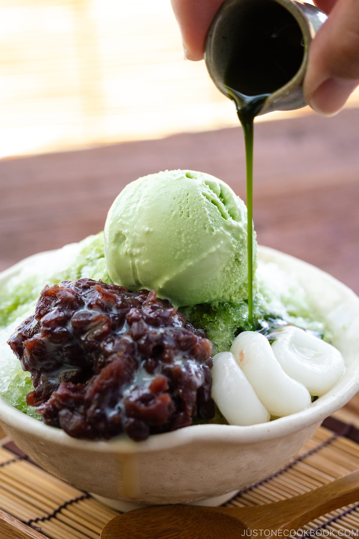 A bowl containing shaved ice, drizzled with matcha syrup and topped with red bean paste, shiratama dango, sweetened condensed milk, and green tea ice cream.