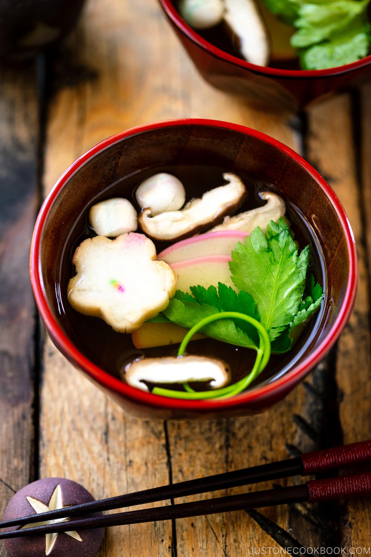 Red Japanese lacquered bowls containing clear soup (Osumashi) with shiitake mushrooms, fu, and mitsuba leaf.