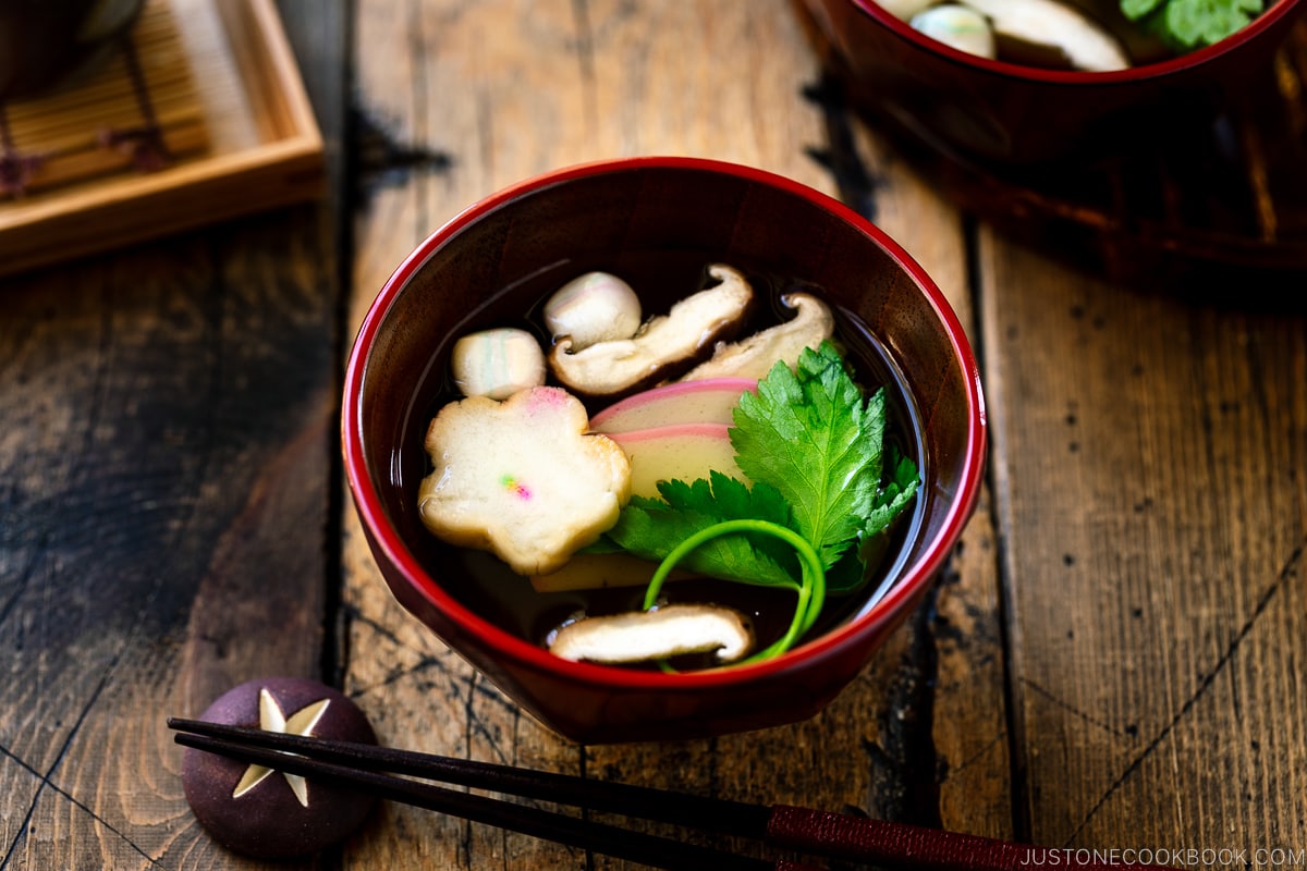 Red Japanese lacquered bowls containing clear soup (Osumashi) with shiitake mushrooms, fu, and mitsuba leaf.