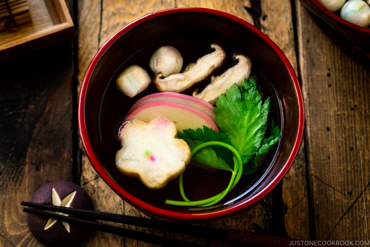 Red Japanese lacquered bowls containing clear soup (Osumashi) with shiitake mushrooms, fu, and mitsuba leaf.