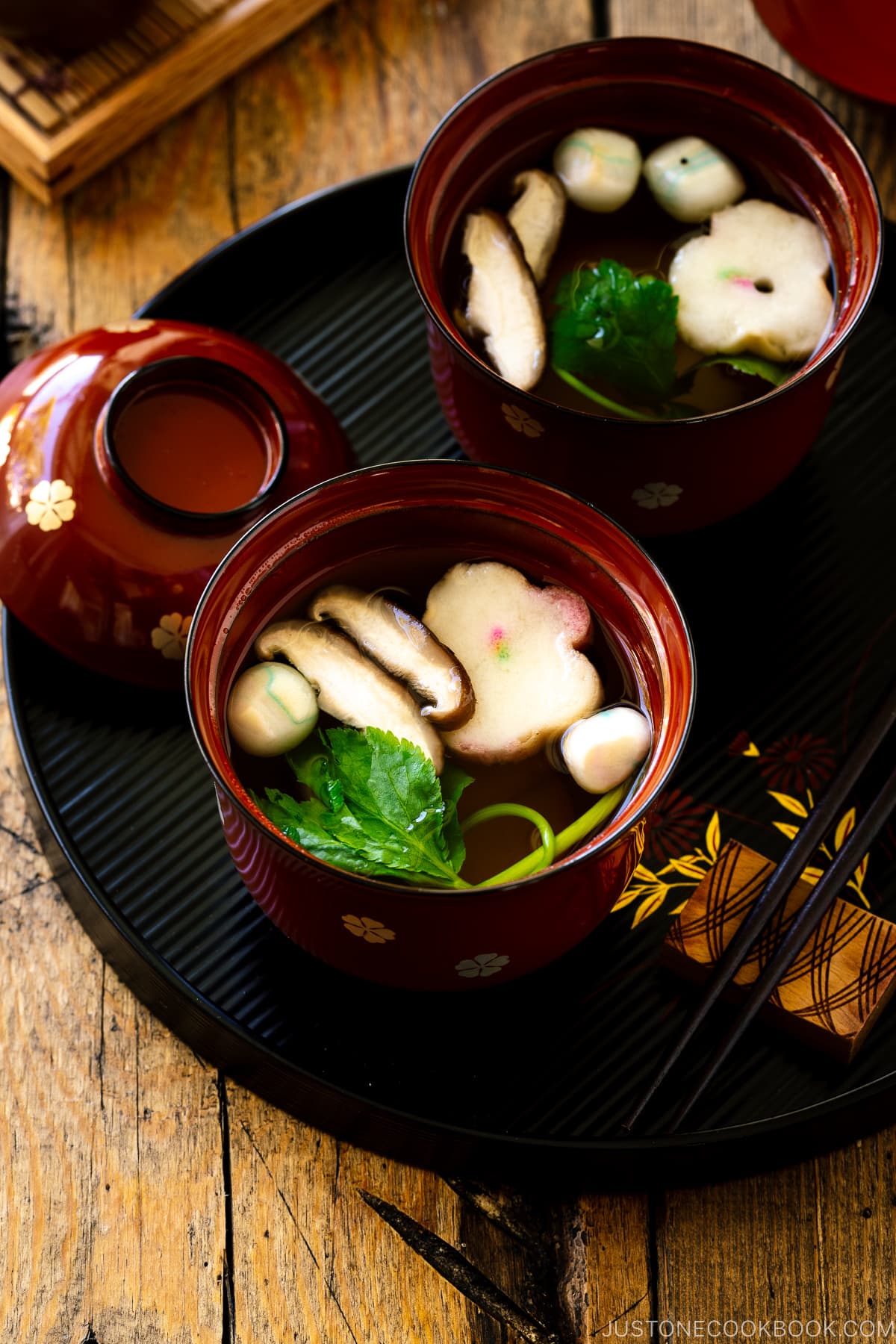Red Japanese lacquered bowls containing clear soup (Osumashi) with shiitake mushrooms, fu, and mitsuba leaf.