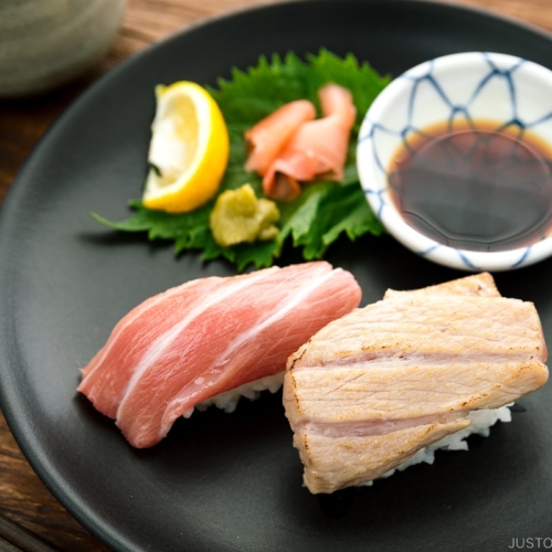 A black plate containing otoro sushi two ways, served with soy sauce, wasabi, and sushi ginger.