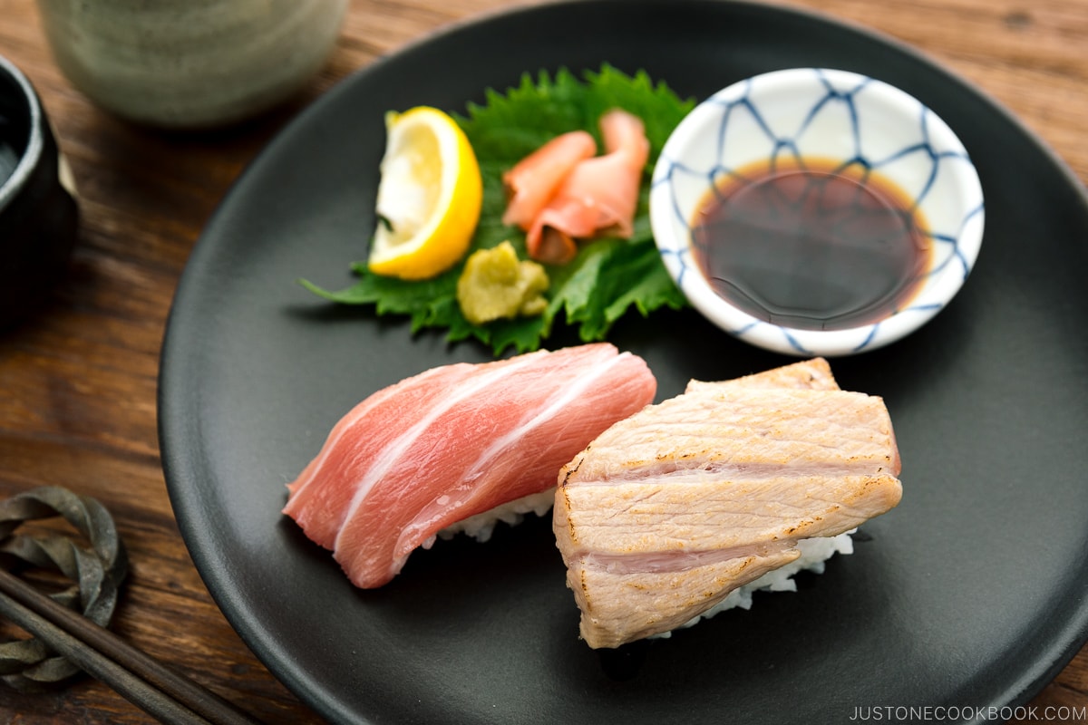 A black plate containing otoro sushi two ways, served with soy sauce, wasabi, and sushi ginger.