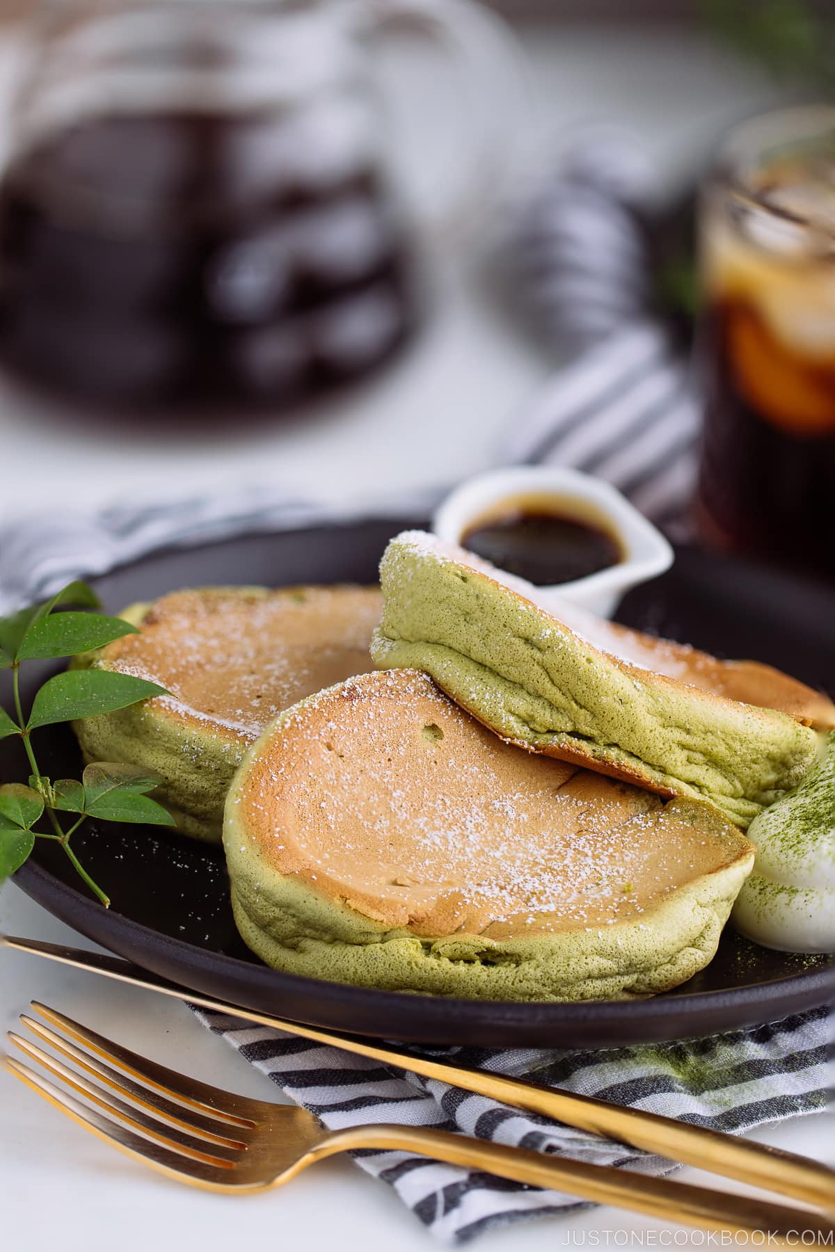 A black plate containing 3 matcha souffle pancakes with fresh whipped cream and maple syrup on the side.