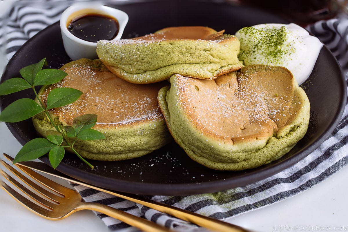 A black plate containing 3 matcha souffle pancakes with fresh whipped cream and maple syrup on the side.