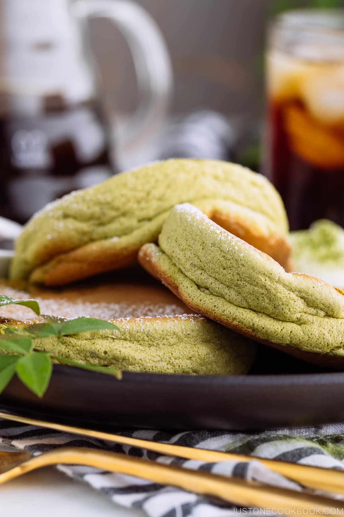 A black plate containing 3 matcha souffle pancakes with fresh whipped cream and maple syrup on the side.