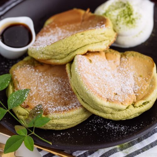 A black plate containing 3 matcha souffle pancakes with fresh whipped cream and maple syrup on the side.