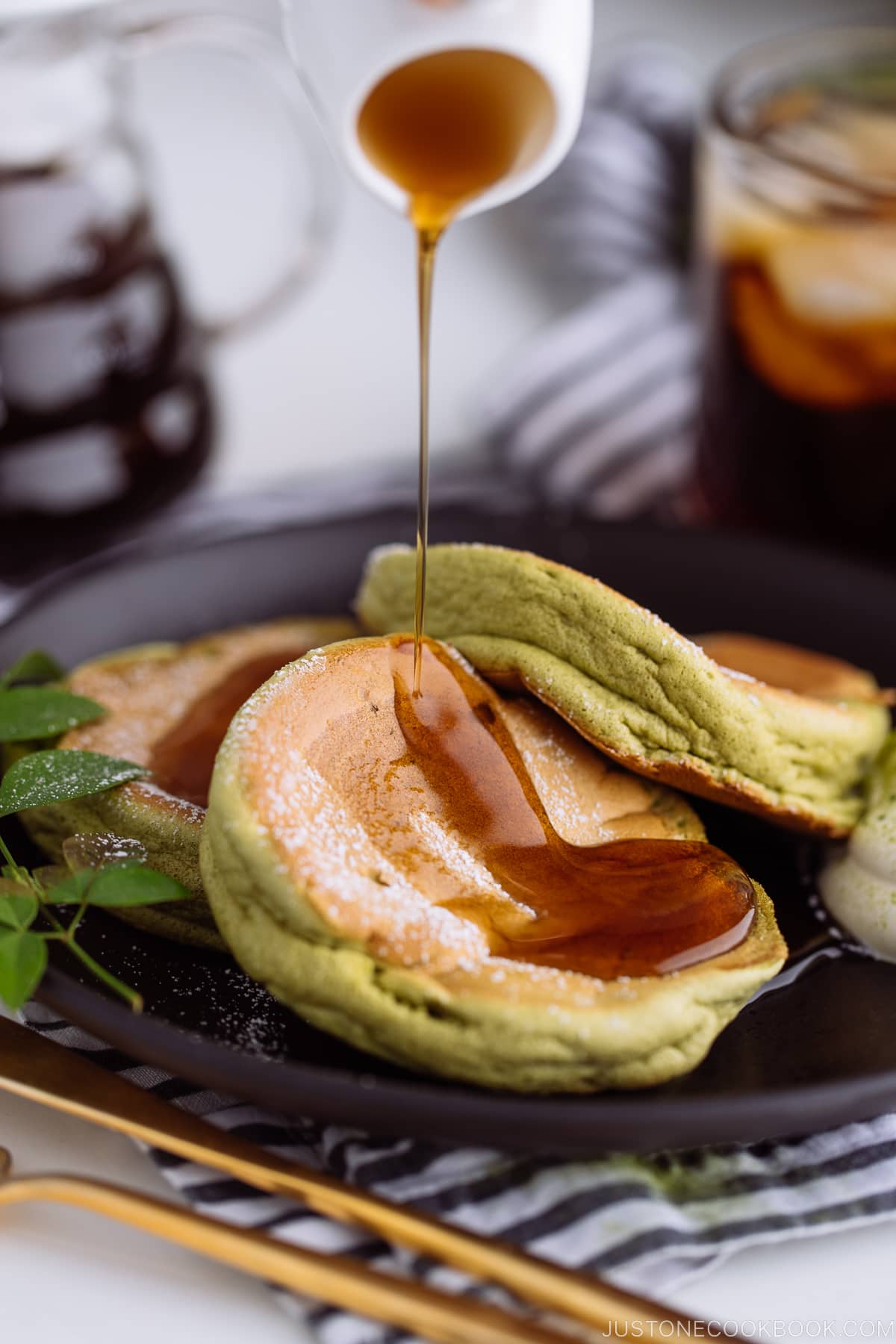 A black plate containing 3 matcha souffle pancakes with fresh whipped cream and maple syrup on the side.