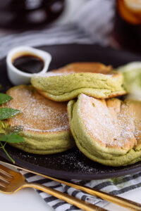 A black plate containing 3 matcha souffle pancakes with fresh whipped cream and maple syrup on the side.