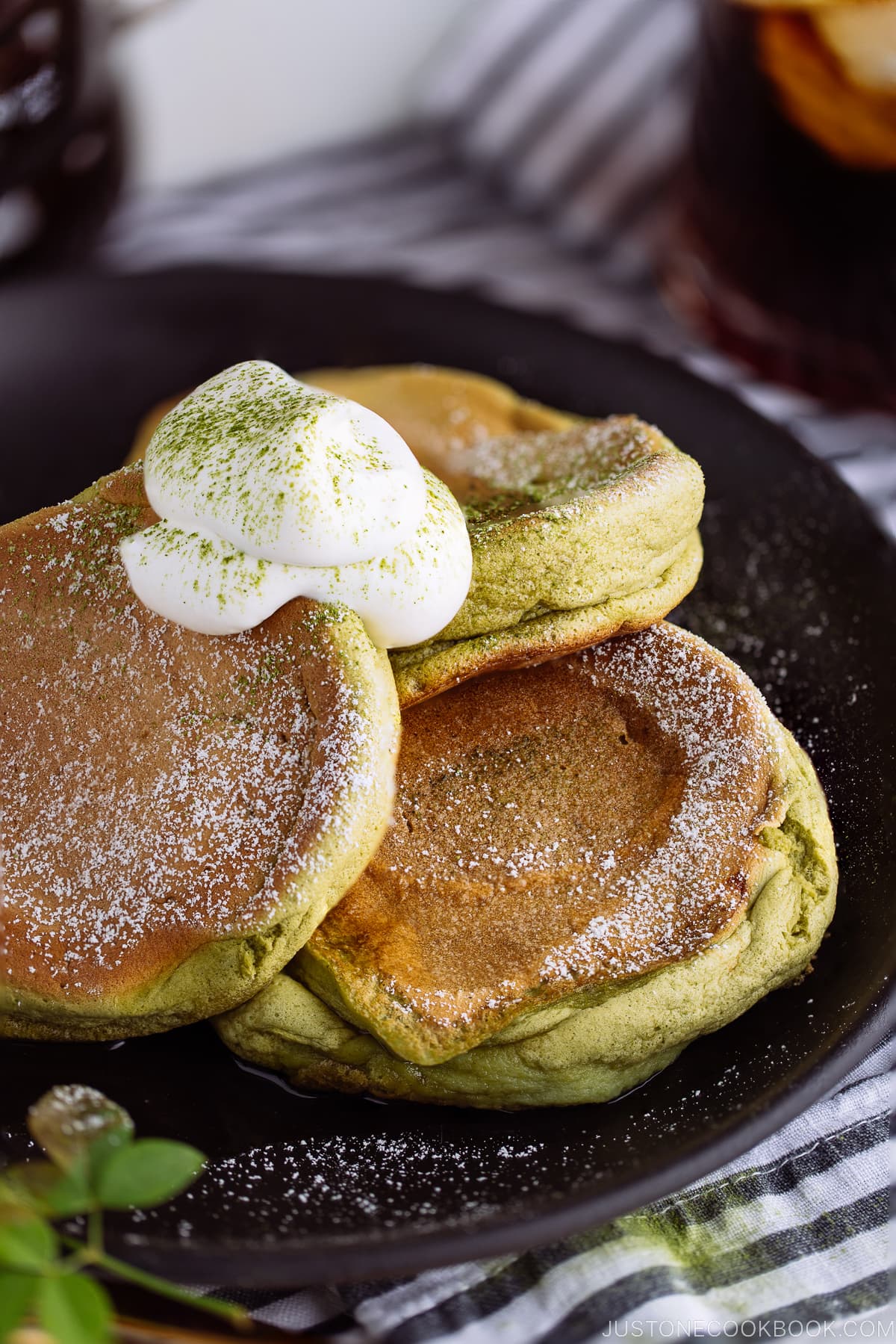 A black plate containing 3 matcha souffle pancakes with fresh whipped cream and maple syrup on the side.