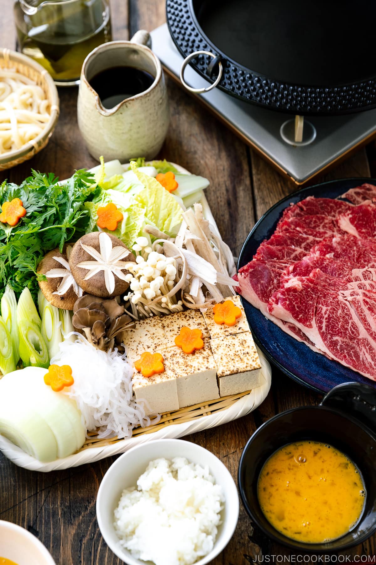 Japanese round cast iron pan, a plate of Japanese beef slices, and a bamboo basket of various vegetables, tofu, and mushrooms.a Japanese hot pot dish where marbled beef, tofu, and vegetables are simmered in sweetened soy sauce broth.