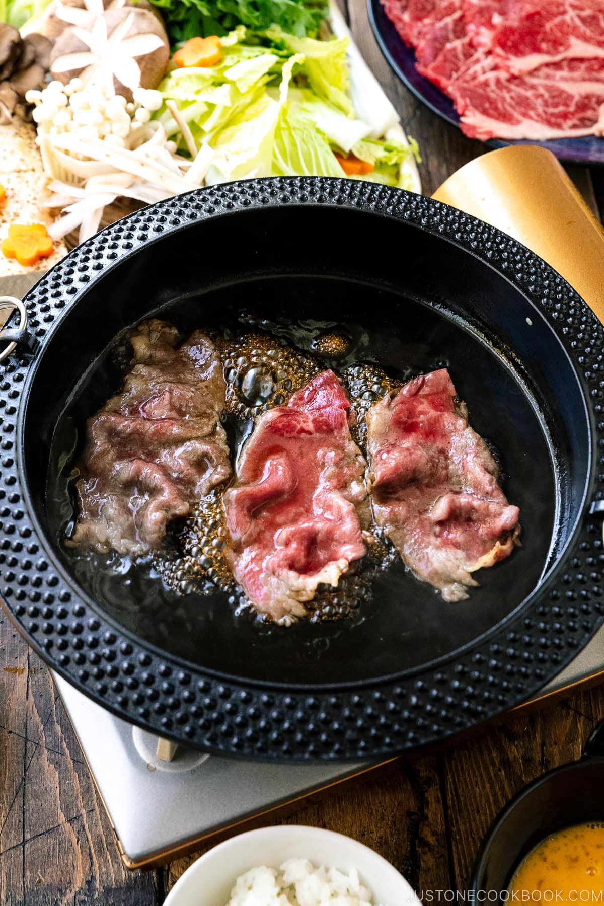 Japanese round cast iron pan containing marbled Sukiyaki beef slices simmered in sweetened soy sauce broth.