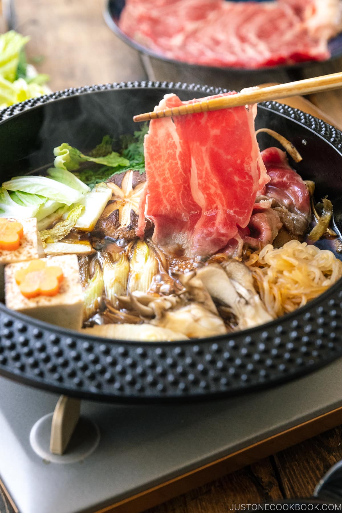 Japanese round cast iron pan containing Sukiyaki, a Japanese hot pot dish where marbled beef, tofu, and vegetables are simmered in sweetened soy sauce broth.