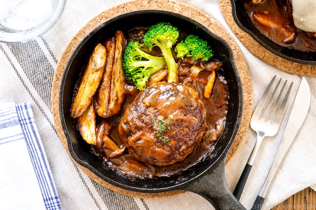 A mini cast iron skillet containing Japanese stewed hamburger steak called Nikomi Hambagu, along with baked potato fries and steamed broccoli.