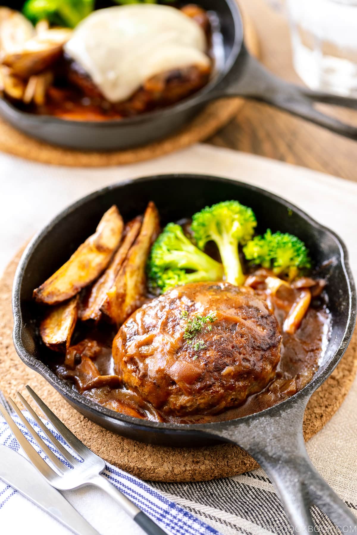 A mini cast iron skillet containing Japanese stewed hamburger steak called Nikomi Hambagu, along with baked potato fries and steamed broccoli.