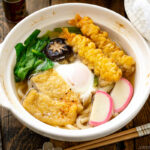 A donabe claypot containing Nabeyaki Udon, which is made of udon noodles, kamaboko fish cake, fried tofu, egg cooked in a dashi broth and topped with shrimp tempura.