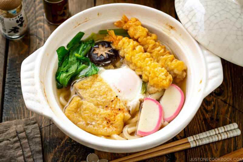 A donabe claypot containing Nabeyaki Udon, which is made of udon noodles, kamaboko fish cake, fried tofu, egg cooked in a dashi broth and topped with shrimp tempura.