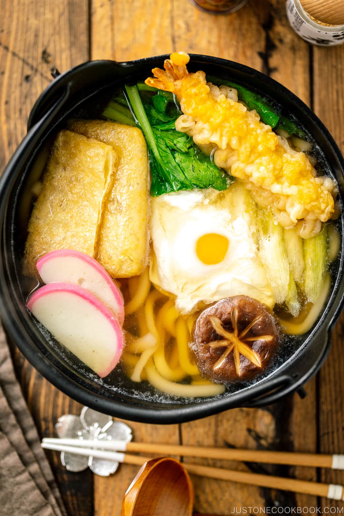 An individual cast iron pot containing Nabeyaki Udon, which is made of udon noodles, kamaboko fish cake, fried tofu, egg cooked in a dashi broth and topped with shrimp tempura.