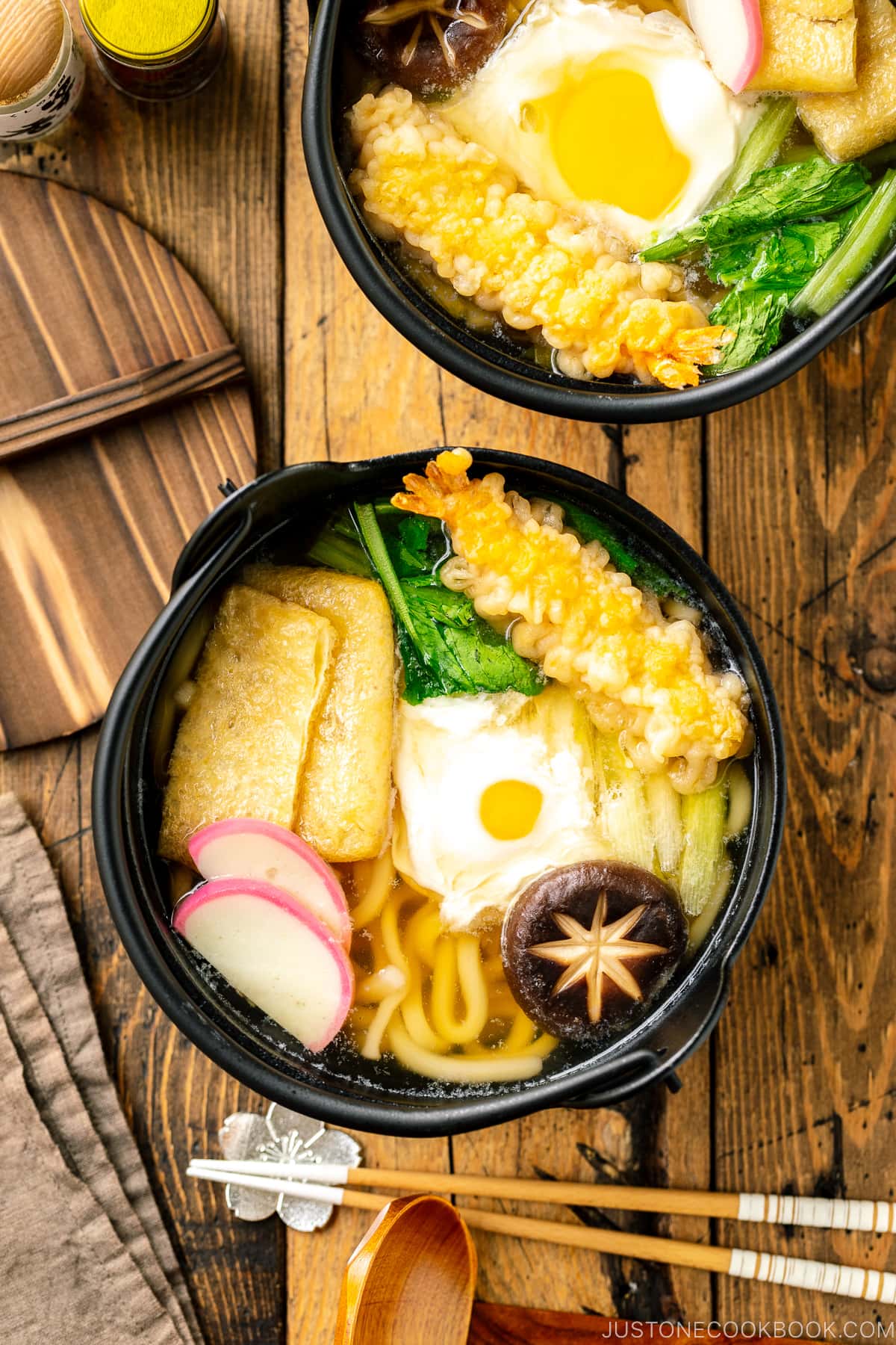 An individual cast iron pot containing Nabeyaki Udon, which is made of udon noodles, kamaboko fish cake, fried tofu, egg cooked in a dashi broth and topped with shrimp tempura.