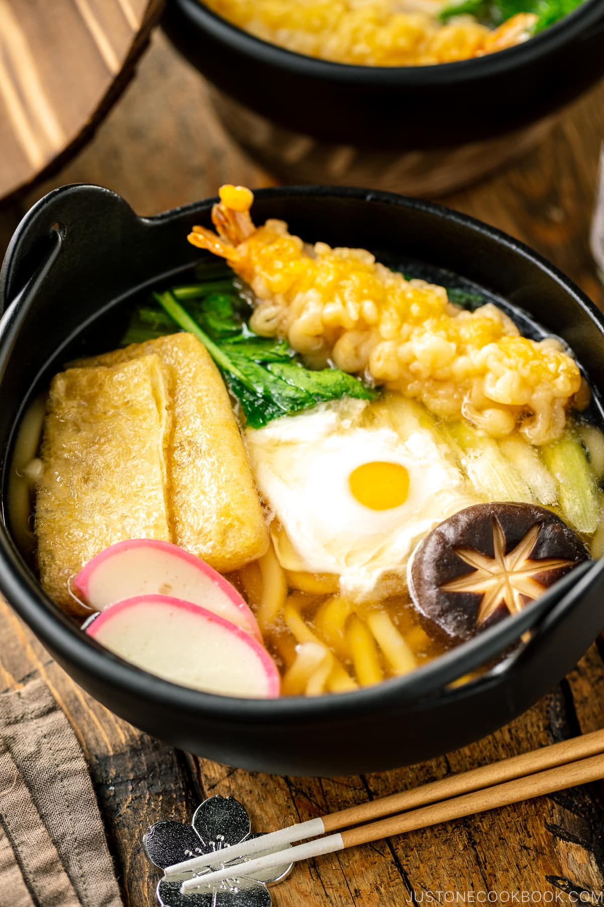 An individual cast iron pot containing Nabeyaki Udon, which is made of udon noodles, kamaboko fish cake, fried tofu, egg cooked in a dashi broth and topped with shrimp tempura.