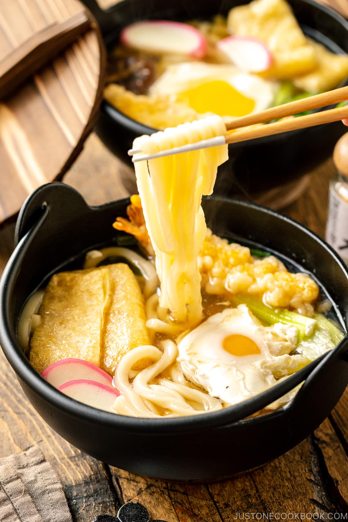 An individual cast iron pot containing Nabeyaki Udon, which is made of udon noodles, kamaboko fish cake, fried tofu, egg cooked in a dashi broth and topped with shrimp tempura.
