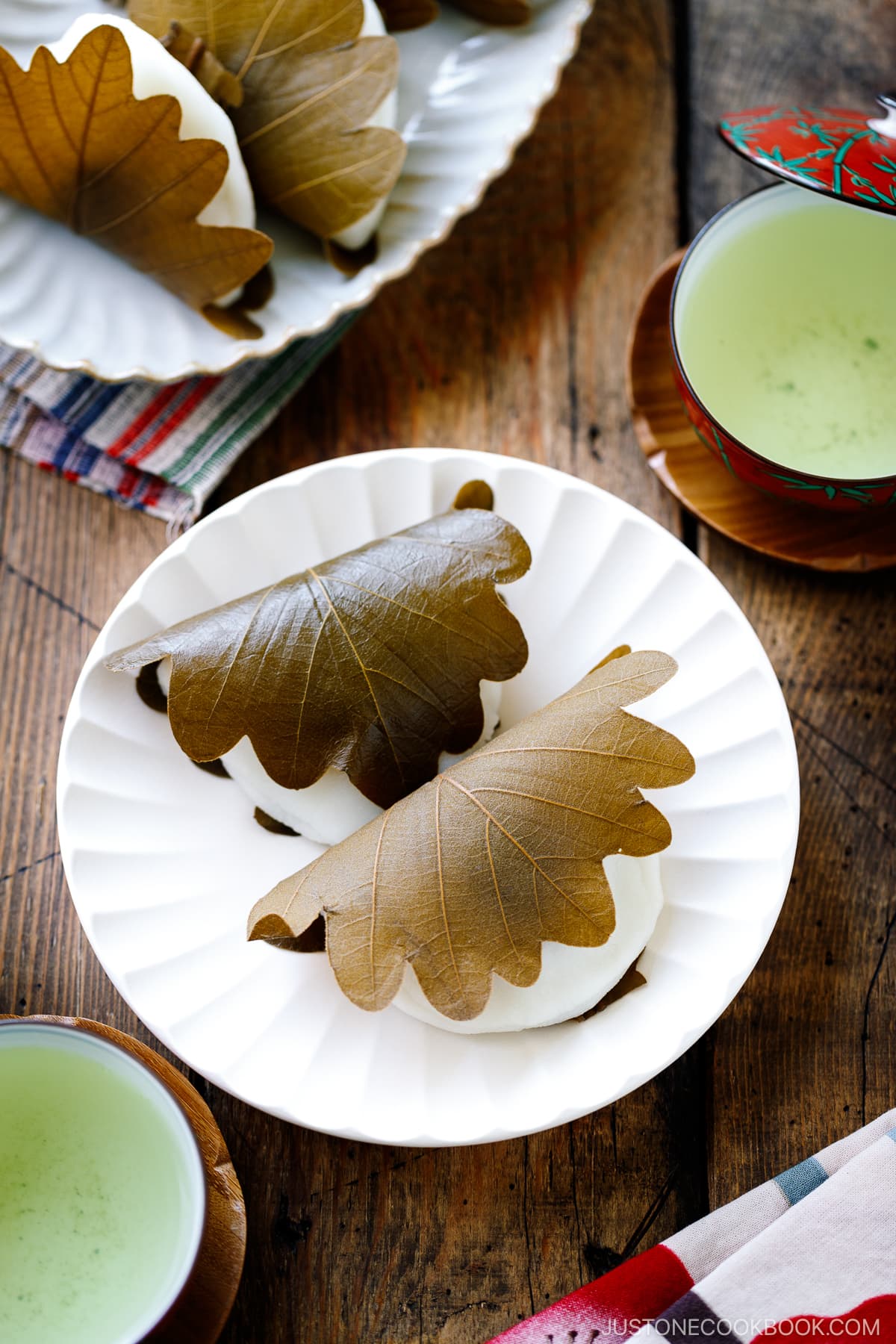 A fluted white plate containing two pieces of Kashiwa Mochi, a Japanese sweet red bean filled mochi wrapped in oak leaf.