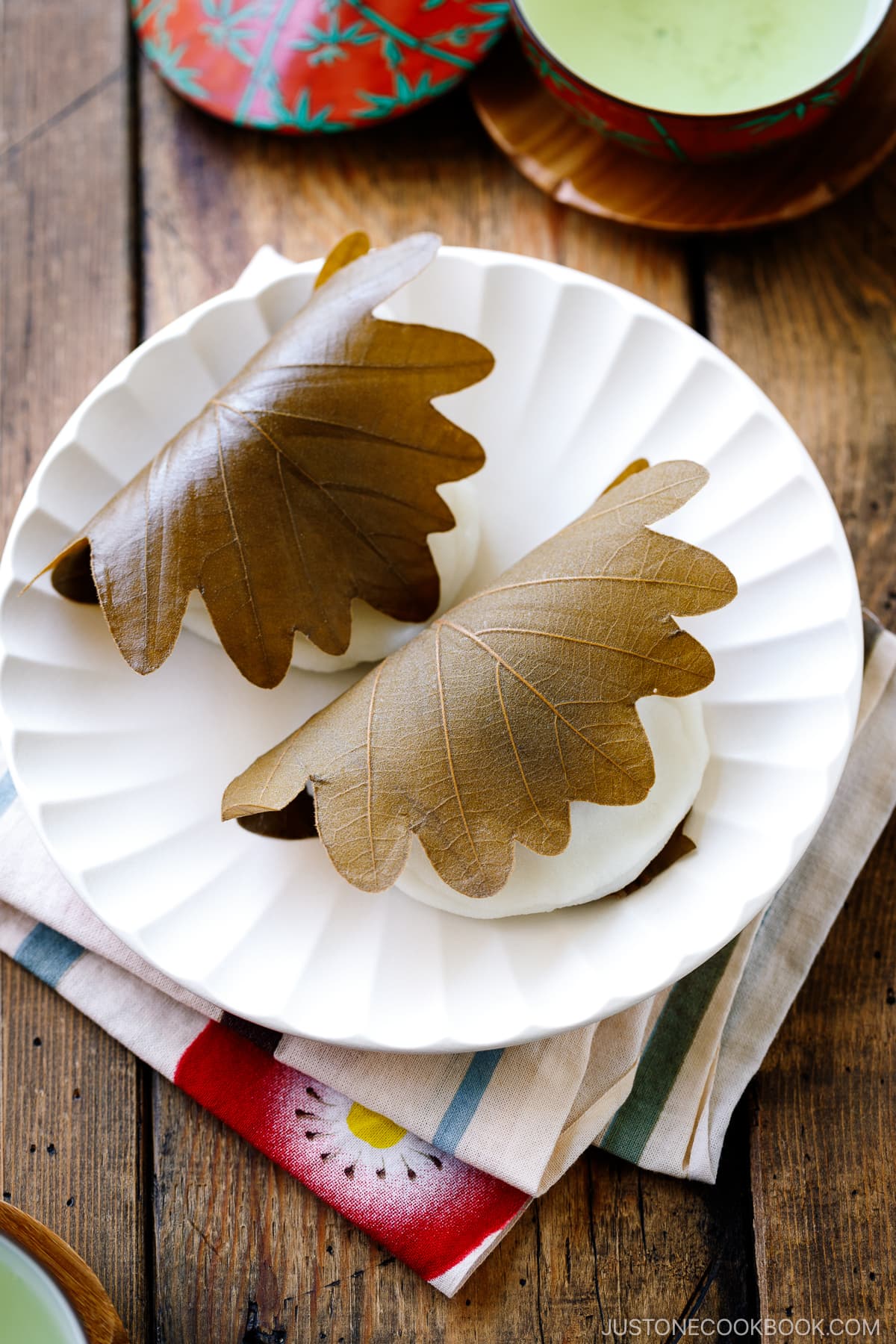 A fluted white plate containing two pieces of Kashiwa Mochi, a Japanese sweet red bean filled mochi wrapped in oak leaf.