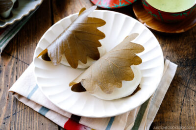 A fluted white plate containing two pieces of Kashiwa Mochi, a Japanese sweet red bean filled mochi wrapped in oak leaf.