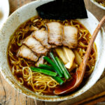 A ramen bowl containing shoyu ramen topped with chashu, seasoned bamboo shoots, green onion, and nori.