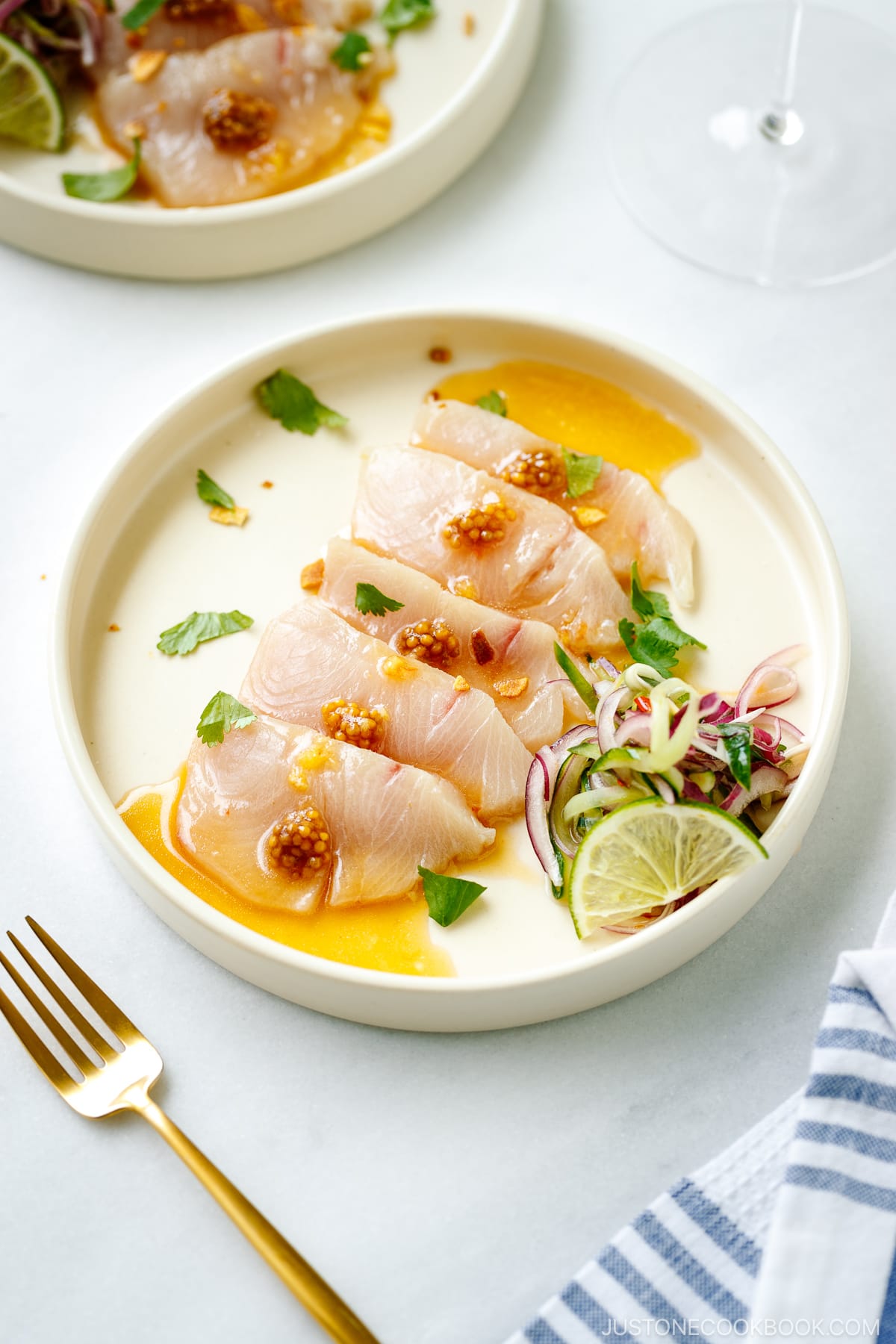 A round plate containing Hamachi Crudo dressed in a bright ponzu and sweet chili sauce vinaigrette, topped with a quick-pickled red onion and cucumber garnish.