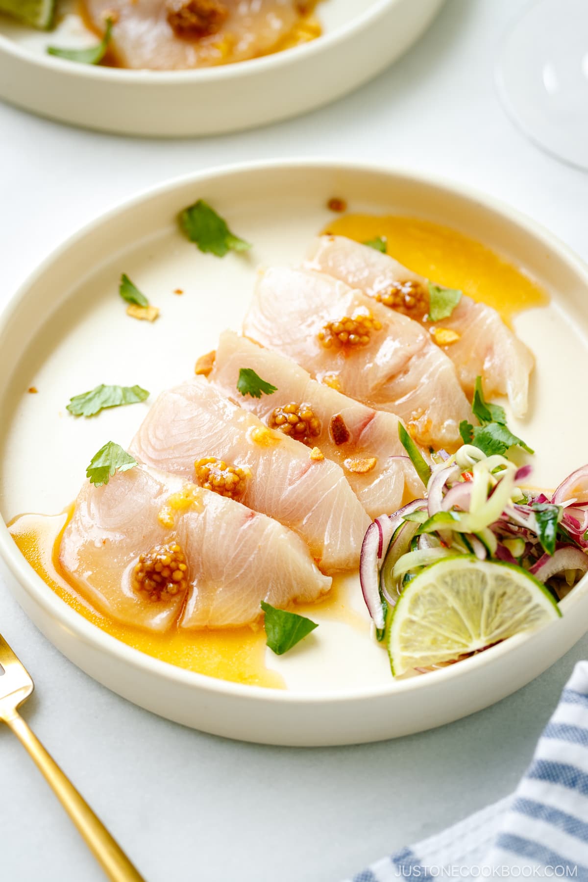 A round plate containing Hamachi Crudo dressed in a bright ponzu and sweet chili sauce vinaigrette, topped with a quick-pickled red onion and cucumber garnish.