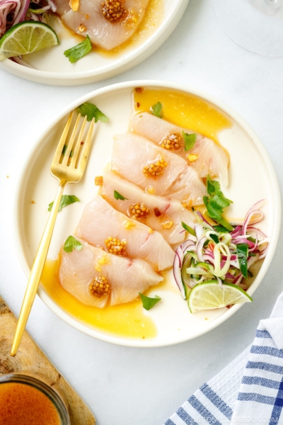 A round plate containing Hamachi Crudo dressed in a bright ponzu and sweet chili sauce vinaigrette, topped with a quick-pickled red onion and cucumber garnish.