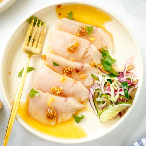 A round plate containing Hamachi Crudo dressed in a bright ponzu and sweet chili sauce vinaigrette, topped with a quick-pickled red onion and cucumber garnish.