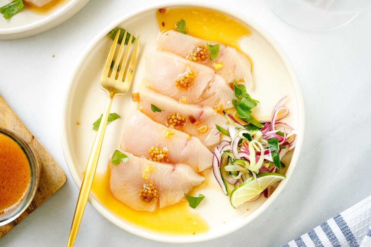 A round plate containing Hamachi Crudo dressed in a bright ponzu and sweet chili sauce vinaigrette, topped with a quick-pickled red onion and cucumber garnish.