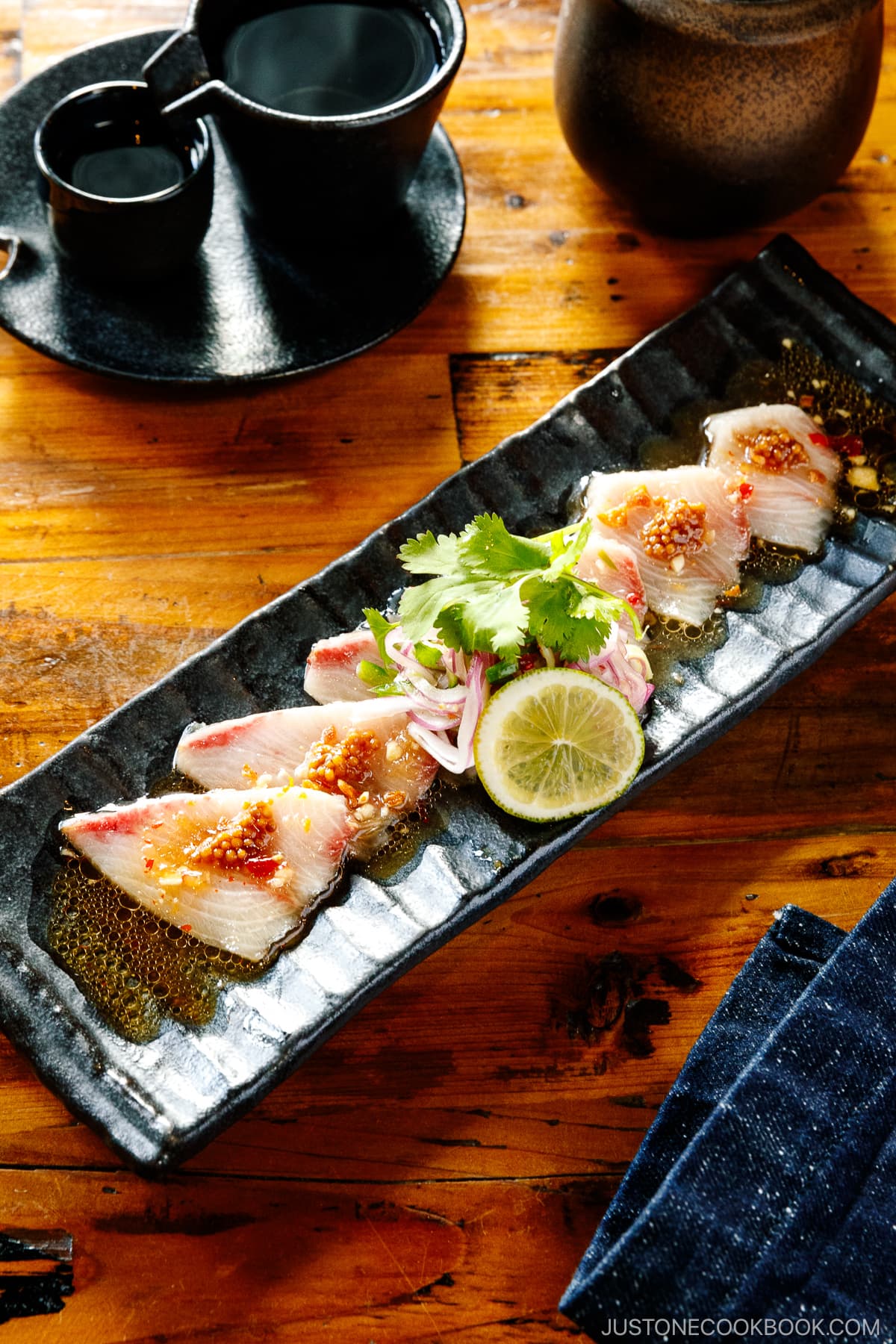A black rectangular plate containing Hamachi Crudo garnished with mustard seeds, garlic chips, cilantro, and a slice of lime
