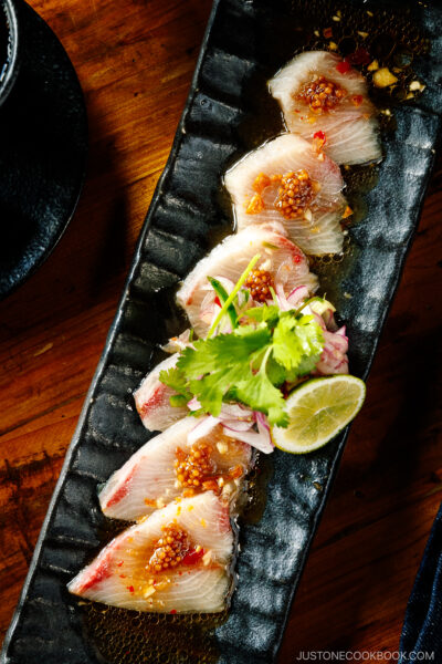 A black rectangular plate containing Hamachi Crudo garnished with mustard seeds, garlic chips, cilantro, and a slice of lime