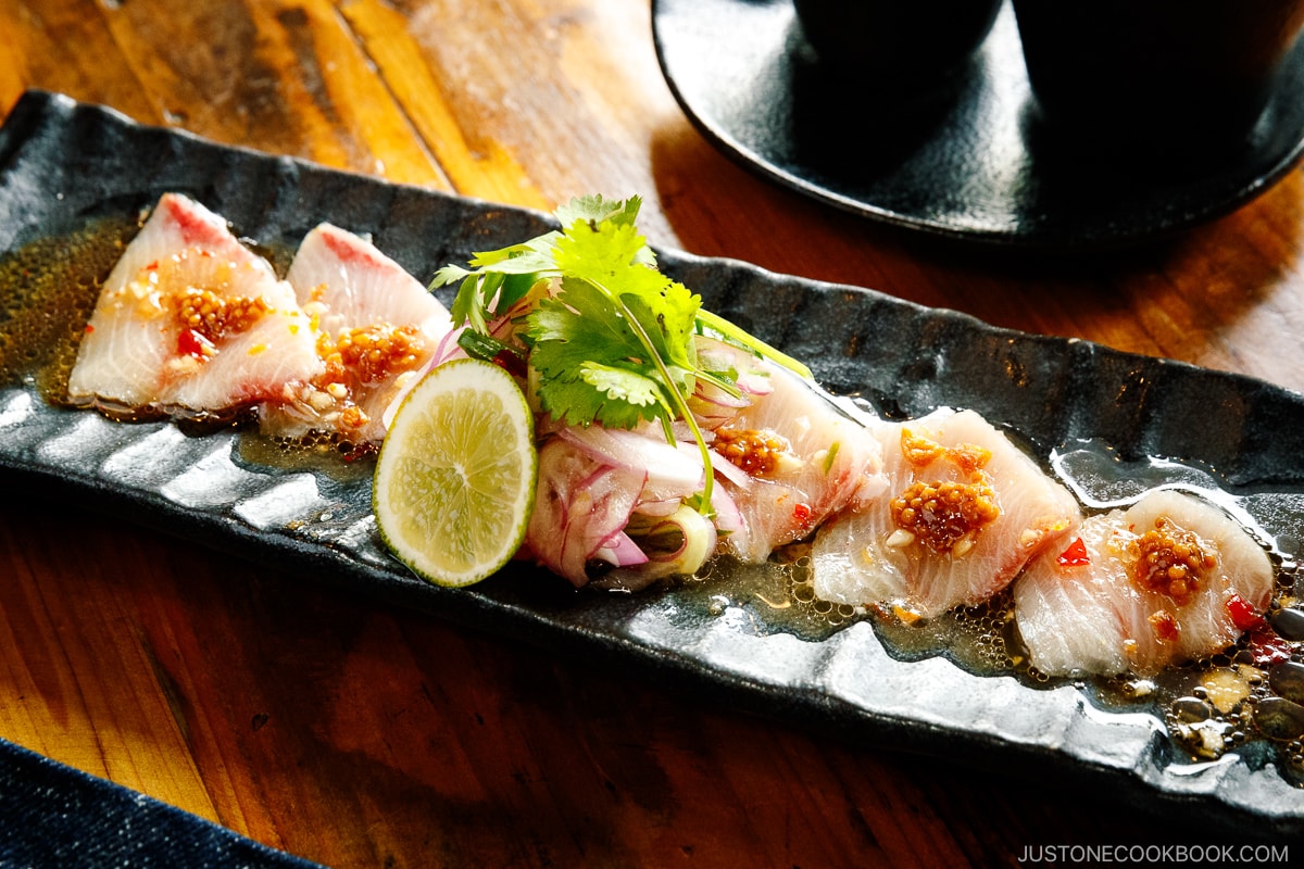 A black rectangular plate containing Hamachi Crudo garnished with mustard seeds, garlic chips, cilantro, and a slice of lime