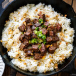 A cast-iron pan containing Steak Garlic Rice topped with chopped scallions and garlic chips.