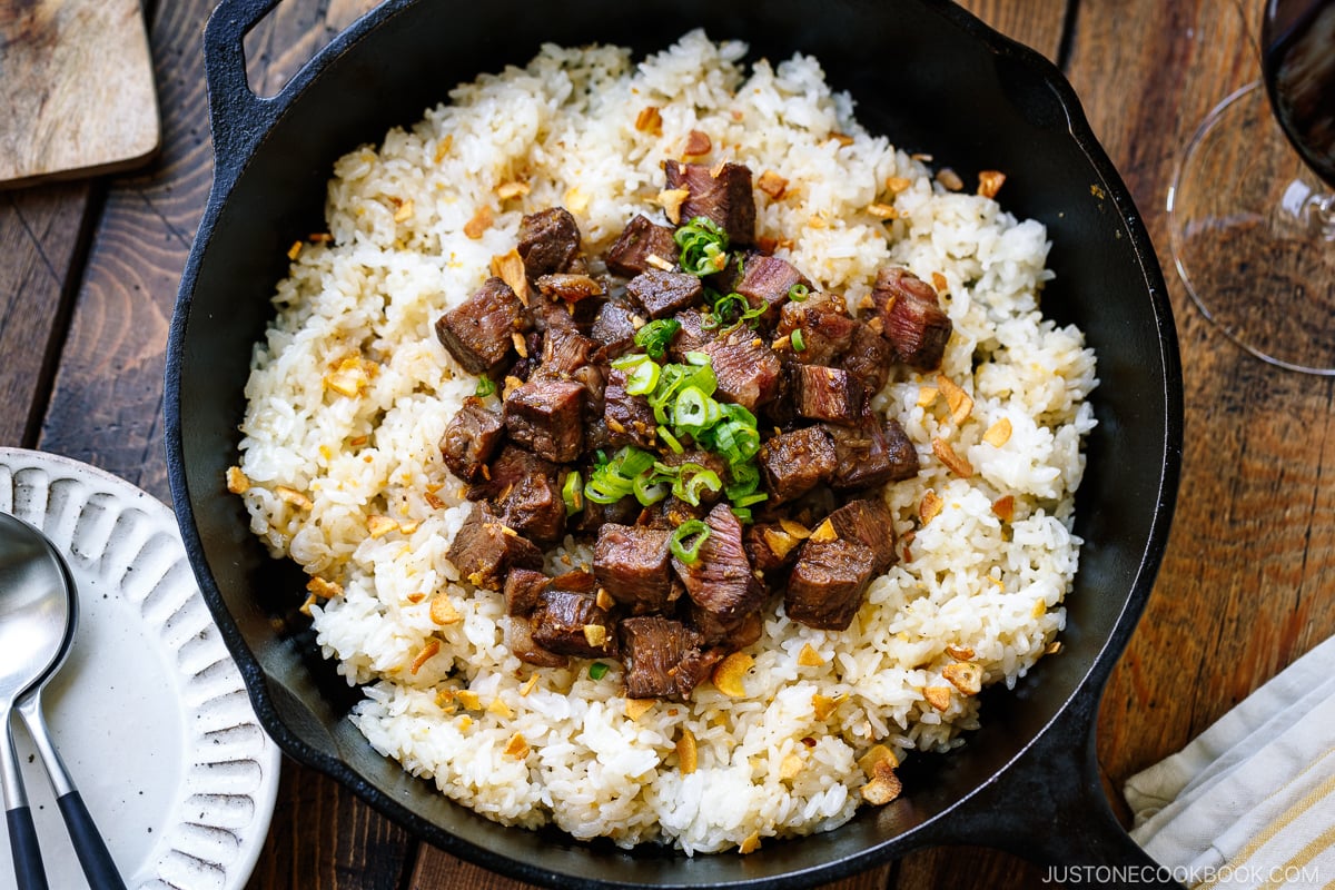 A cast-iron pan containing Steak Garlic Rice topped with chopped scallions and garlic chips.