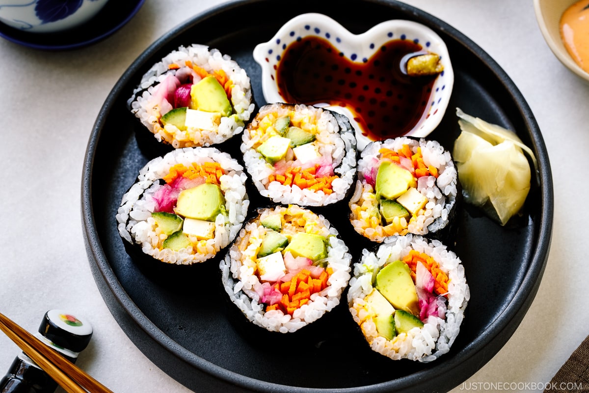A black round plate containing colorful vegetarian sushi rolls, sushi ginger, and soy sauce.