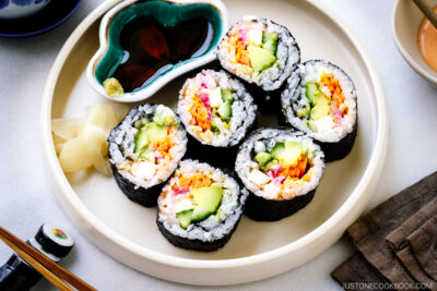 A white round plate containing colorful vegetarian sushi rolls, sushi ginger, and soy sauce.