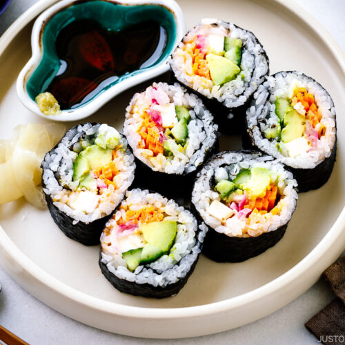 A white round plate containing colorful vegetarian sushi rolls, sushi ginger, and soy sauce.