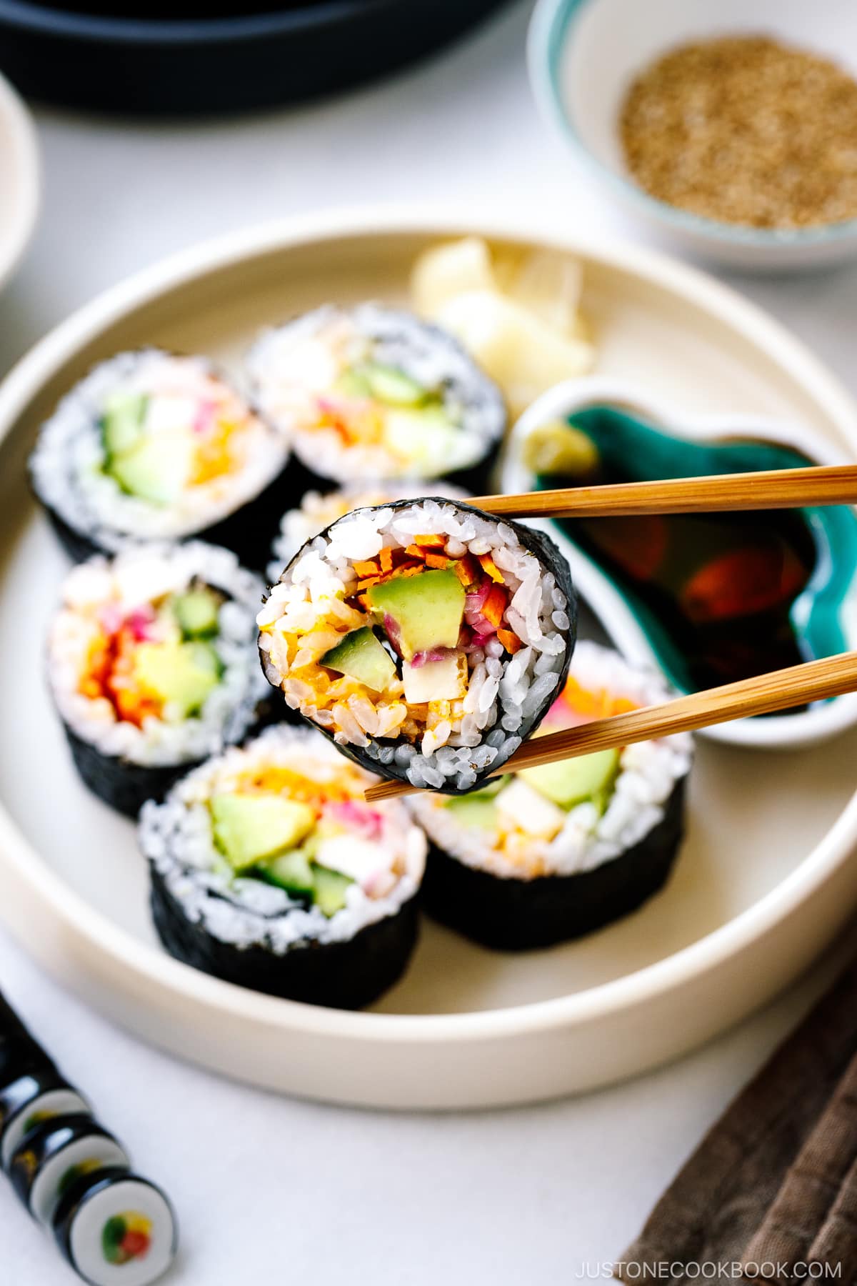 A white round plate containing colorful vegetarian sushi rolls, sushi ginger, and soy sauce.