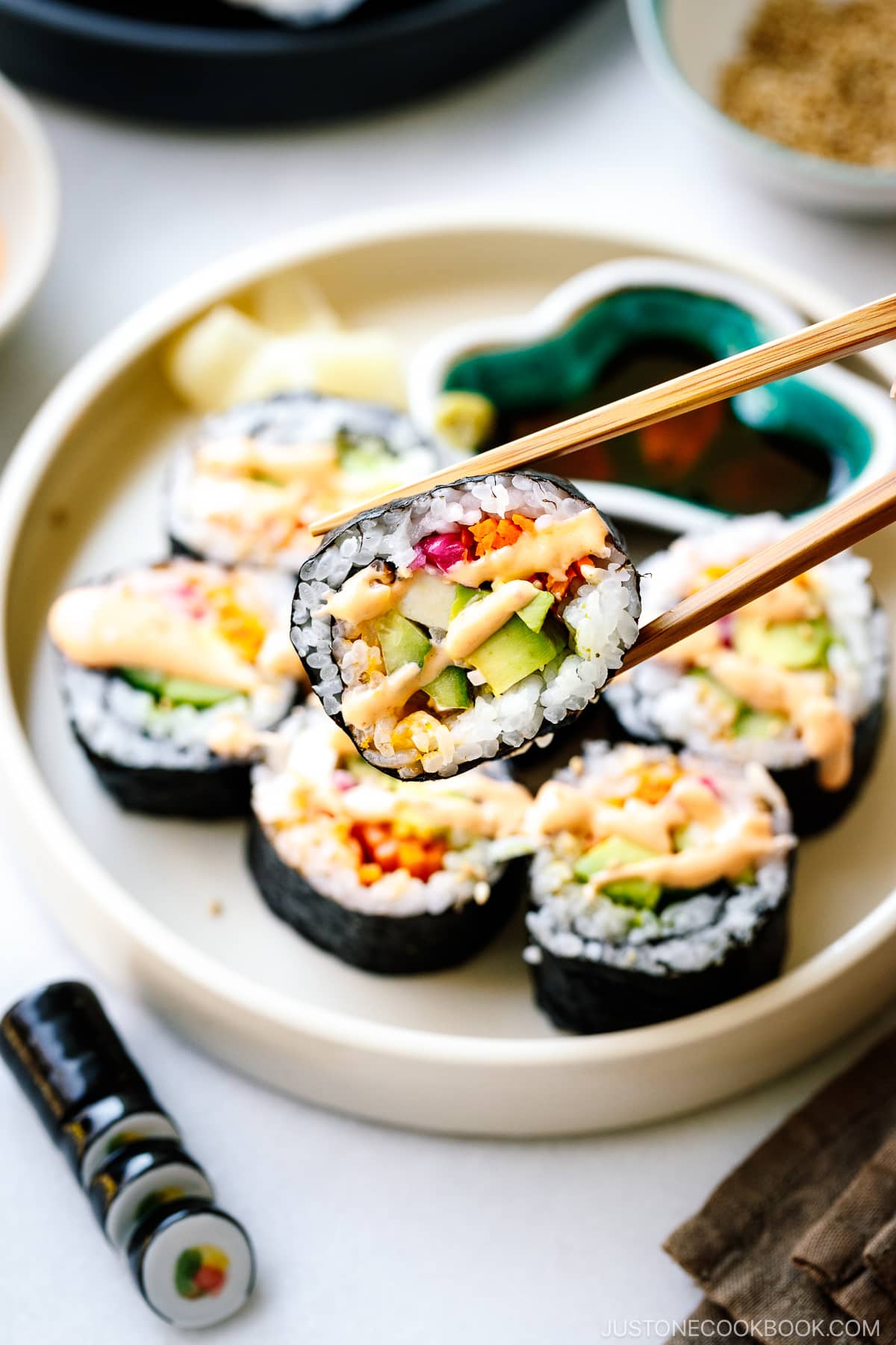 A white round plate containing colorful vegetarian sushi rolls, sushi ginger, and soy sauce.