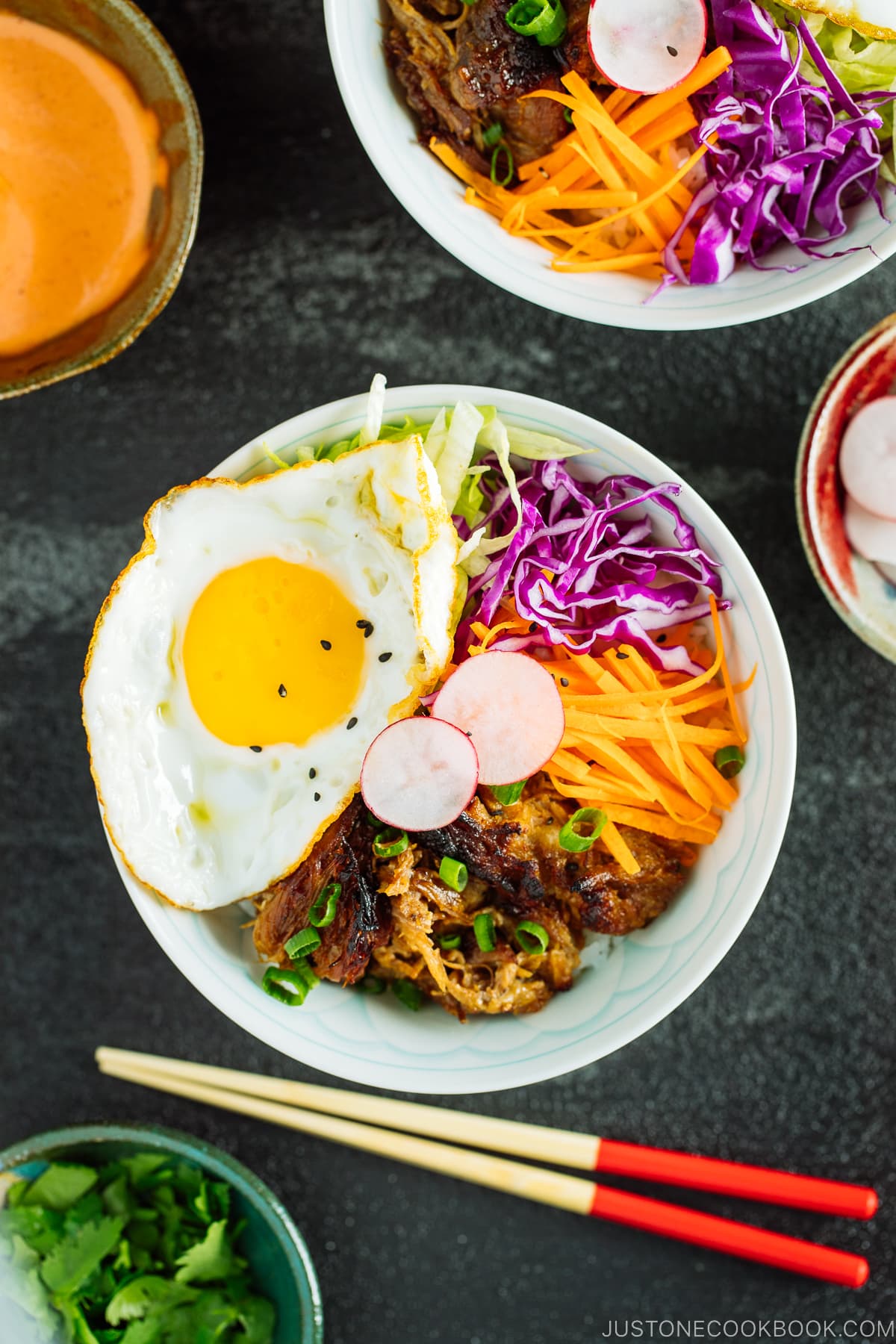 Plates and bowls containing ingredients for Asian Pulled Pork Bowls, including steamed rice, fried eggs, veggies, and spicy mayo.