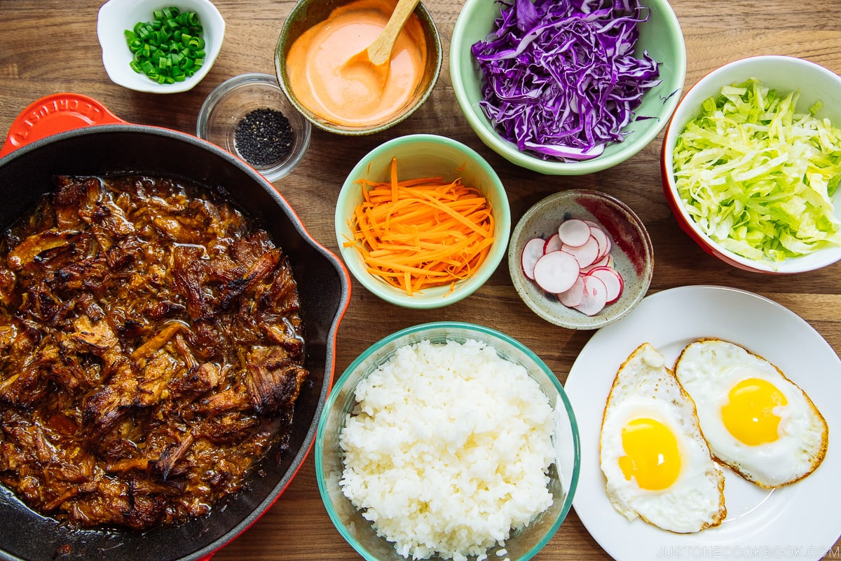 Plates and bowls containing ingredients for Asian Pulled Pork Bowls, including steamed rice, fried eggs, veggies, and spicy mayo.