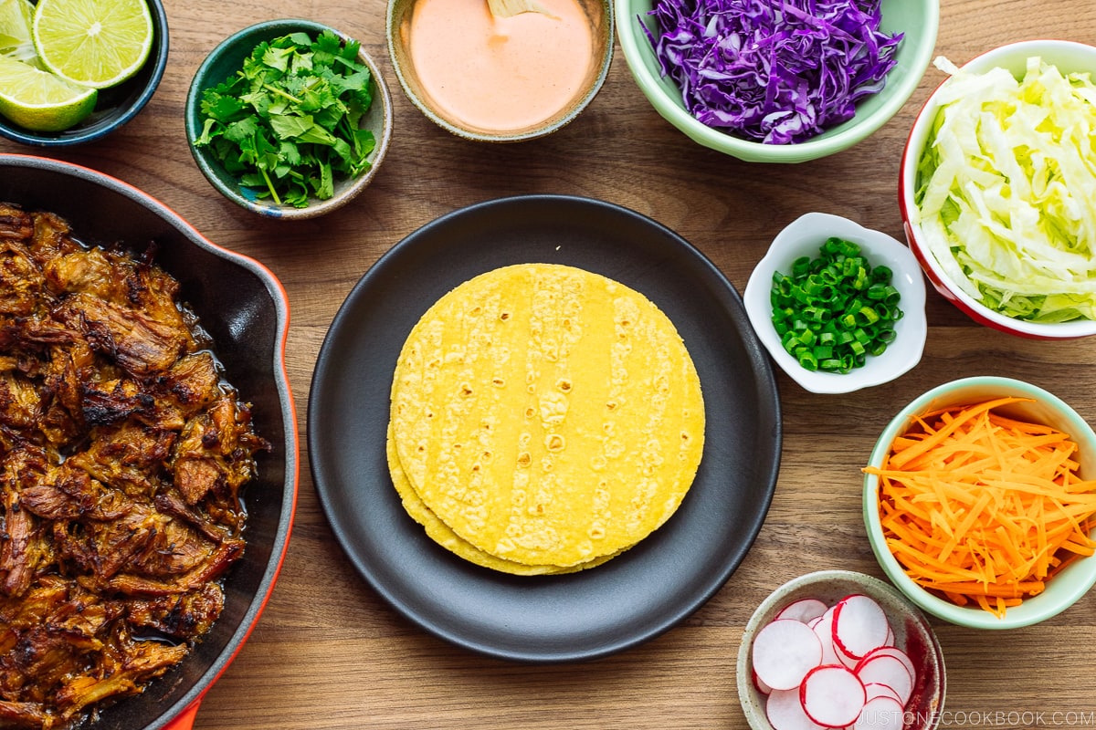 The wooden table with bowls and plates containing Asian Pulled Pork Taco ingredients.