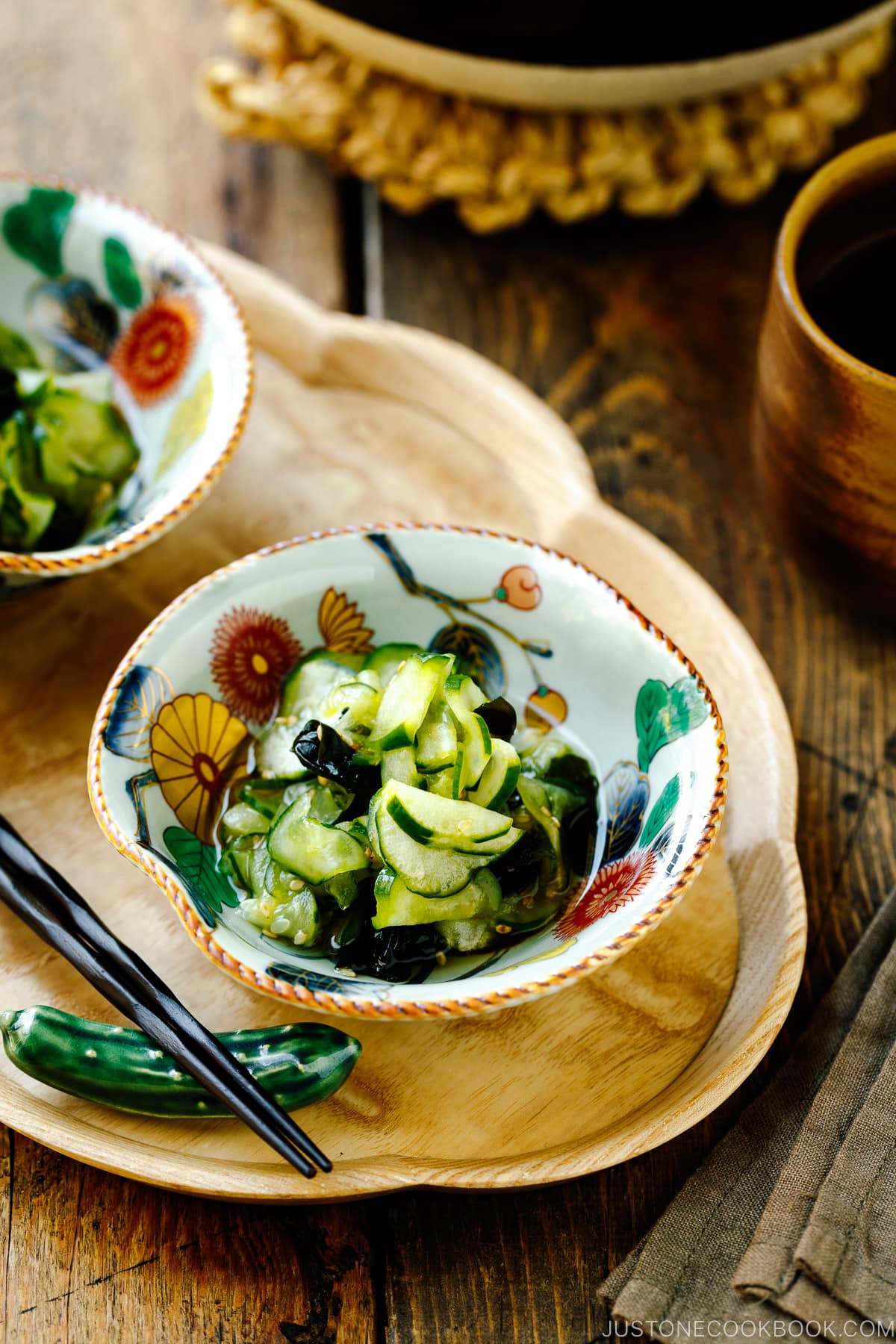 Small Japanese bowls containing Japanese cucumber salad called Sunomono served on top of a wooden tray along with a cucumber shaped chopstick rest and chopsticks.