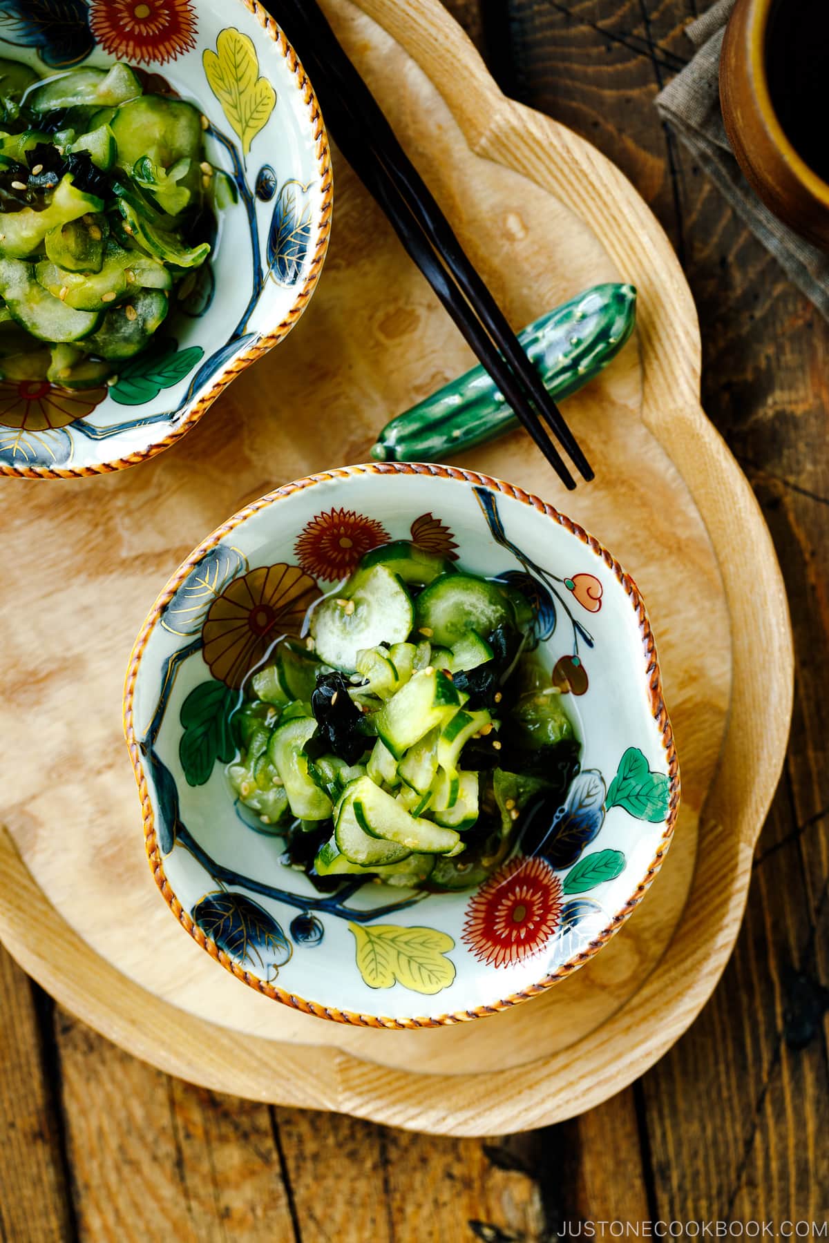 Small Japanese bowls containing Japanese cucumber salad called Sunomono served on top of a wooden tray along with a cucumber shaped chopstick rest and chopsticks.